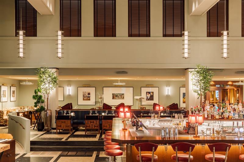 Romslig hotellbar med stor takhøyde, stilfulle barkrakker, dempet belysning og elegante sittegrupper i varme fargetoner.