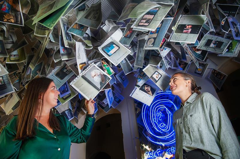 To personer inne i en installasjon med opphengte fotografier, dokumenter og belyste kunstverk.