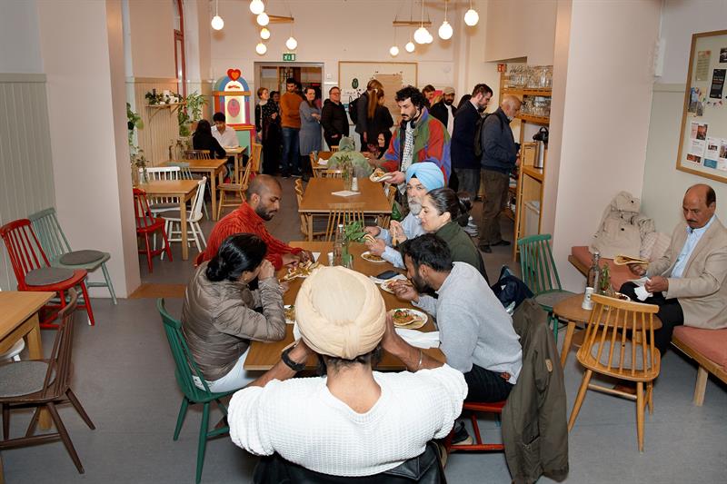Kaféområde med besøkende som sitter ved bordene og folk som står i kø ved serveringsområdet.