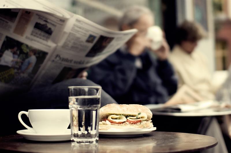 Detaljbilde av en kaffekopp, et glass vann og en ciabatta med en person som leser en avis i bakgrunnen.