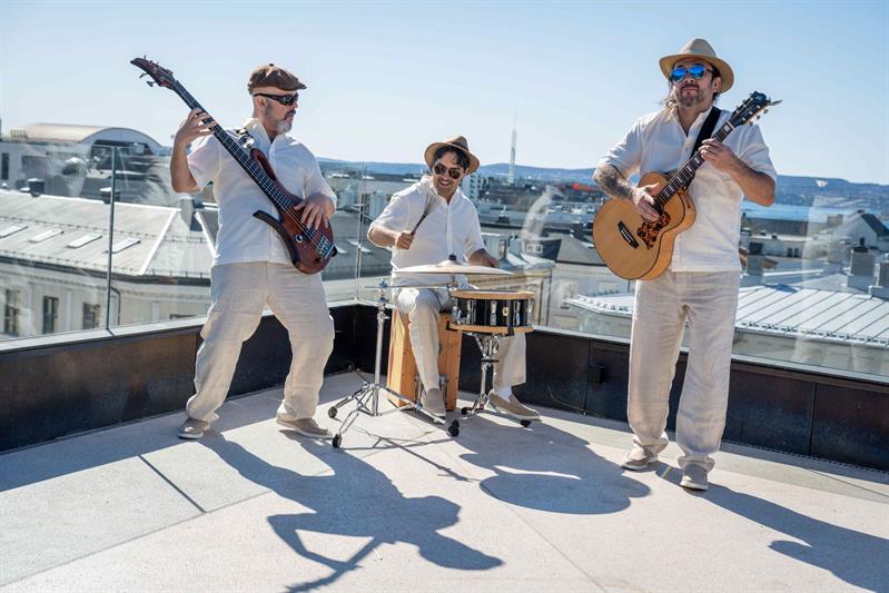 Tre musikere i hvite antrekk opptrer på et hustak og spiller bassgitar, trommer og akustisk gitar mot en naturskjønn bykulisse med en klar