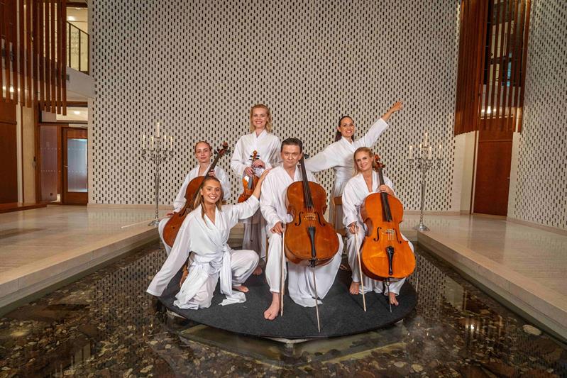 En gruppe musikere i hvite kapper spiller cello og fiolin på en sirkelformet plattform omgitt av vann, med elegante kandelabre i bakgrunnen