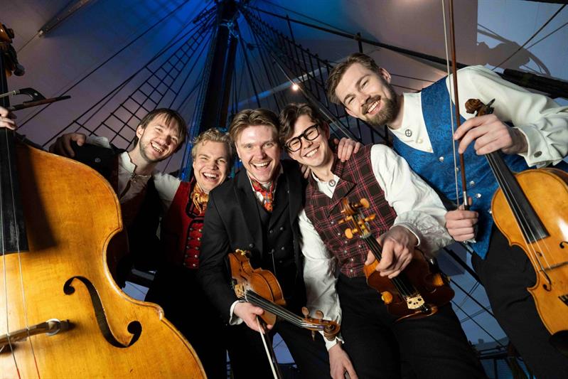 Fire musikere i tradisjonelle klær spiller på strengeinstrumenter, blant annet kontrabass og fioliner, foran en skipsmast som bakteppe.