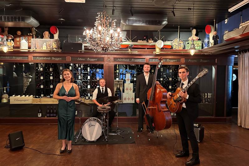 En gruppe musikere opptrer i et koselig rom med tregulv, med en kvinne med mikrofon, en trommeslager, en kontrabassist og en gitarist