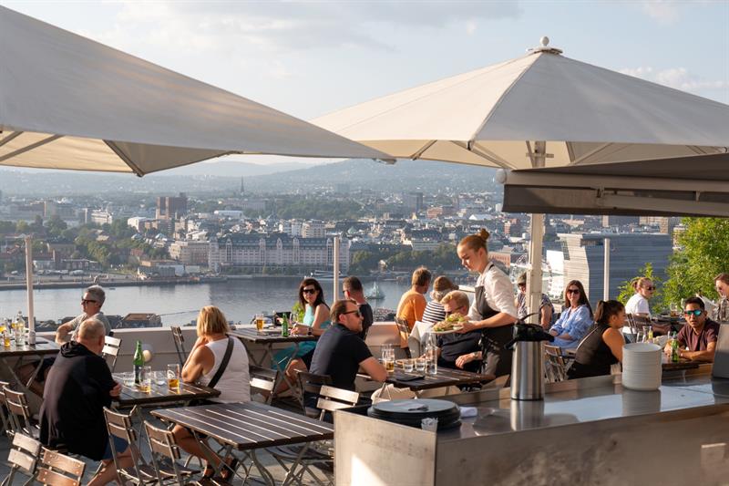 En servitør serverer mat til gjestene på en livlig takterrasse med utsikt over byen.
