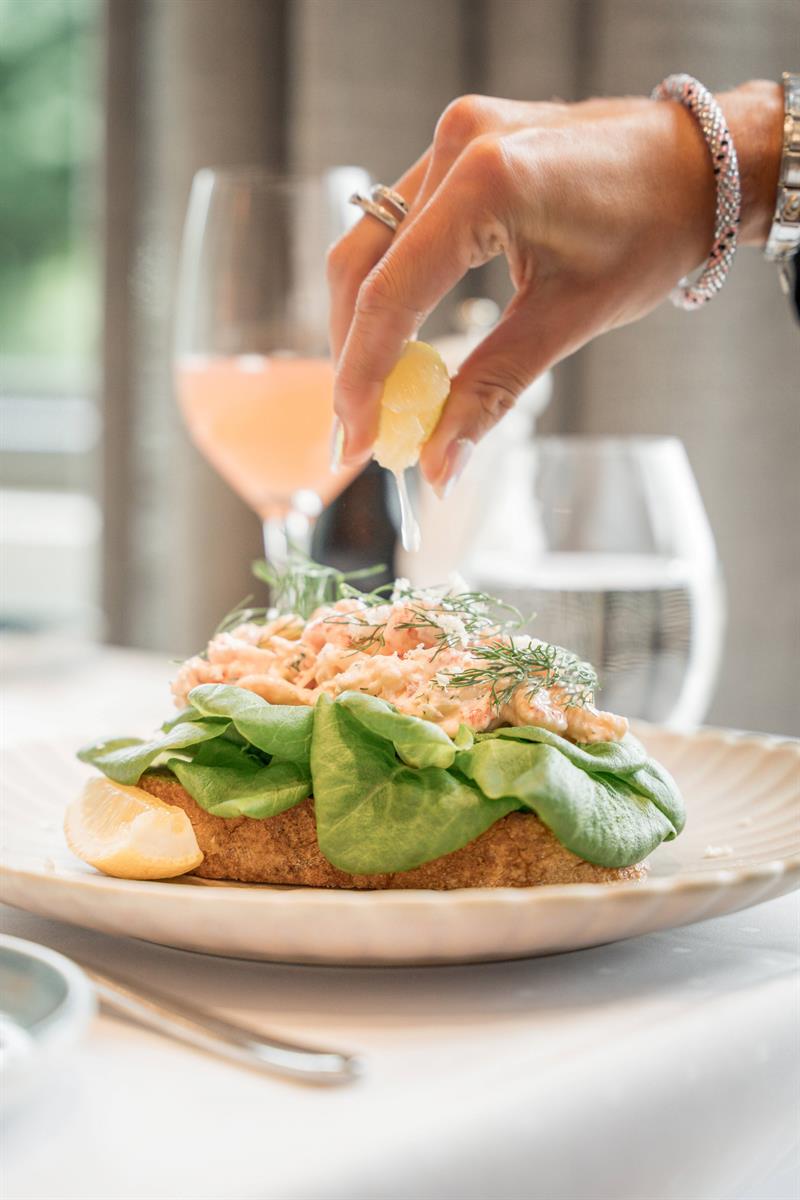 En hånd presser sitron over et rekesmørbrød toppet med friske urter og salatblader.