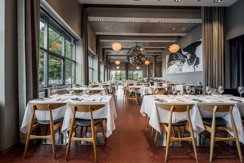 Romslig restaurant med pent oppdekkede bord, designbelysning og utsikt gjennom store vinduer.