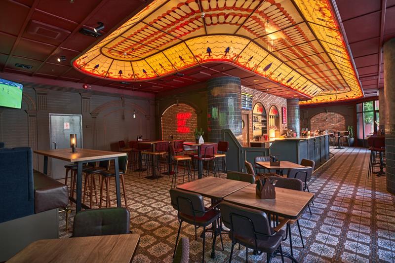Restaurantinteriør med små firkantede bord og stoler, bar og dekorert glasstak over baren