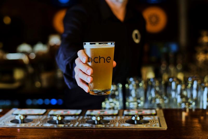 Detalj av en hånd som holder et ølglass med Oche-logoen