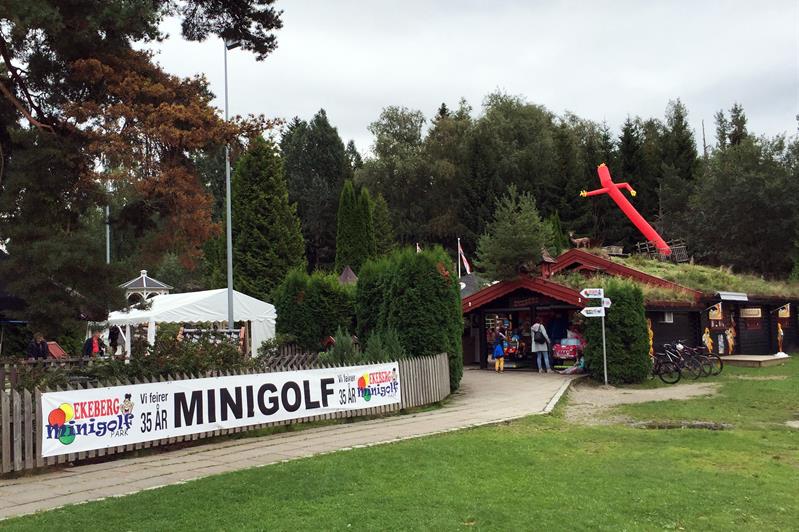 Minigolfbane med en rød rørmann, et par små hus med gress på taket, et gjerde med et skilt på, trær og gress.