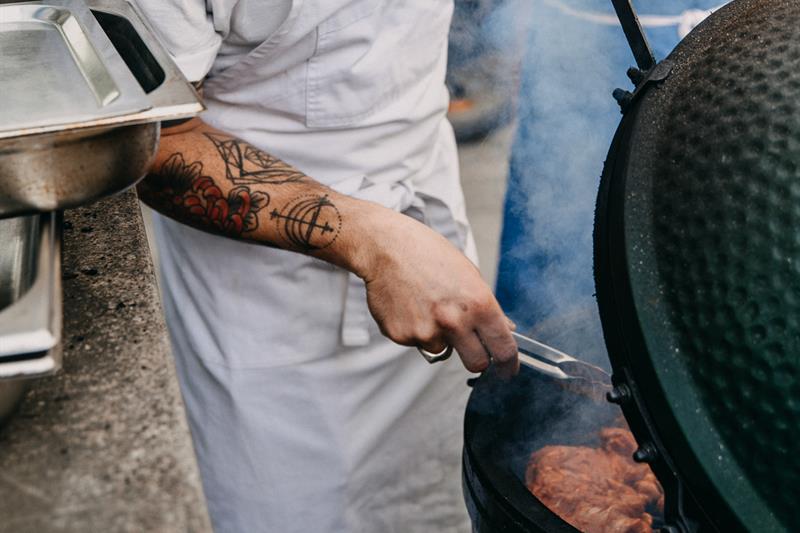 En kokk med en hatt med "breddos" skrevet på forsiden vender kjøtt på en rund grill.