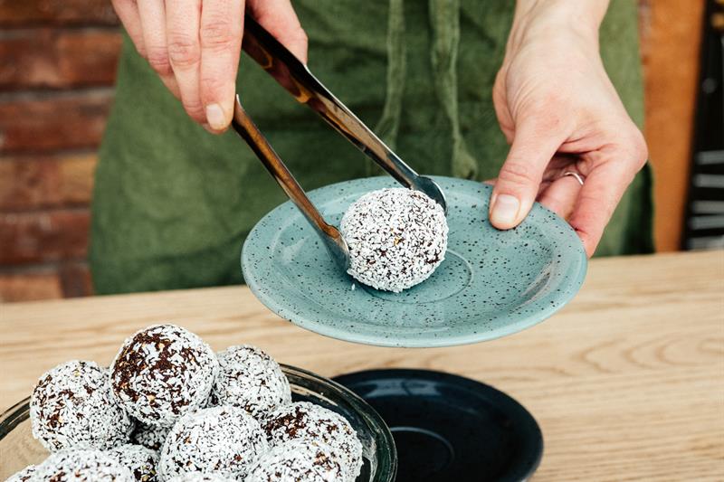 En person i grønt forkle overfører kokosboller fra en full oppbevaringsbolle i glass til et lite fat