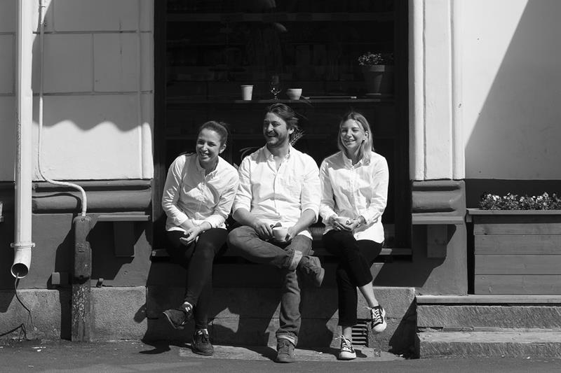 Svart-hvitt bilde. 3 smilende mennesker sitter utenfor en kafé.