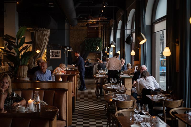 Restaurantinteriør med store panoramavinduer, rutete gulv og små bord og stoler, der folk sitter og spiser