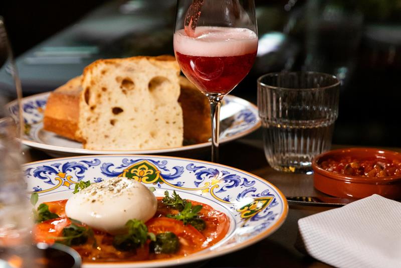 En flaske som skjenker en rosa drikk i et vinglass, over et bord med burrata på tomat, brød, små tallerkener og et glass vann.