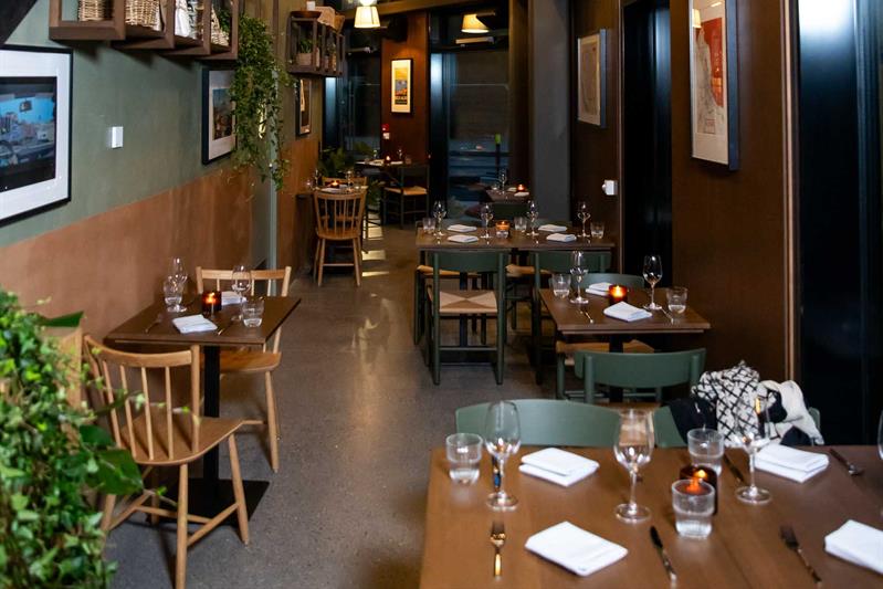 Restaurantinteriør med høyt tak, små trebord dekket til servering og stoler