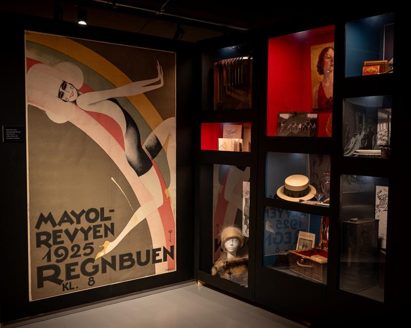 Utstillingsrom med en stor plakat fra Mayol-revyen 1925 omgitt av montrere med fotografier, hatter og andre gjenstander fra mellomkrigstiden.
