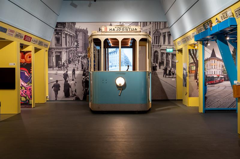 Museumsutstilling med en restaurert trikk foran historiske fotografier av Oslo-gater og informasjon om kollektivtransportens historie.