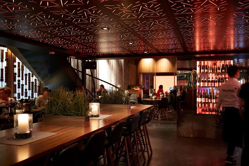 Et restaurantinteriør med et langt bord, stoler, gjester som sitter og spiser ved mindre bord, et kjøleskap med vinflasker og en servitør som går forbi.