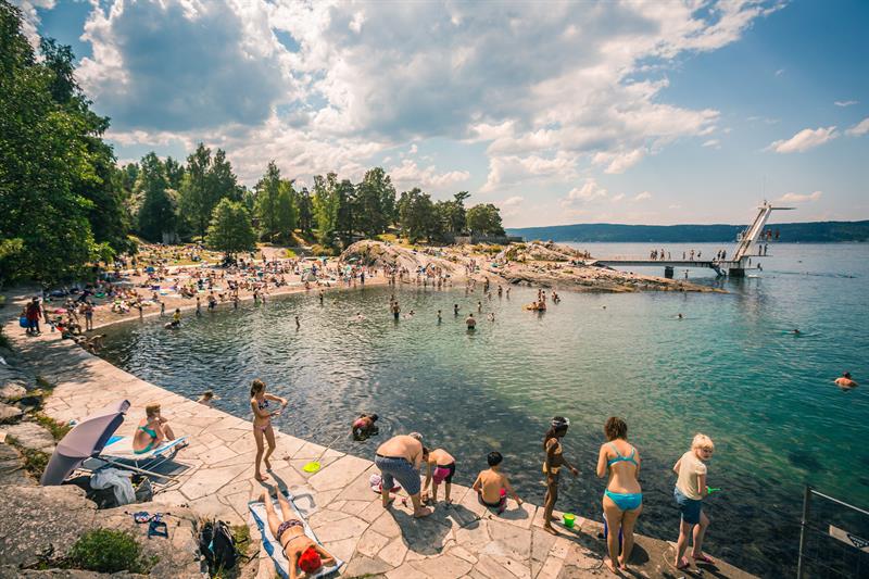 En overfylt strand om sommeren, med folk som hopper fra stupetårnet, svømmer og soler seg på stranden