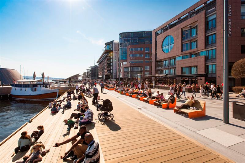 Sol, blå himmel, mennesker som sitter langs kaia, en båt på venstre side, brune bygninger med store vinduer på høyre side. Mennesker som går langs promenaden.