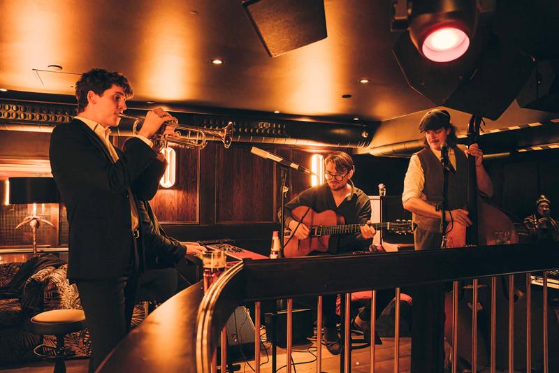 Et band som opptrer på scenen og spiller trompet, gitar og kontrabass, med et rekkverk foran scenen.