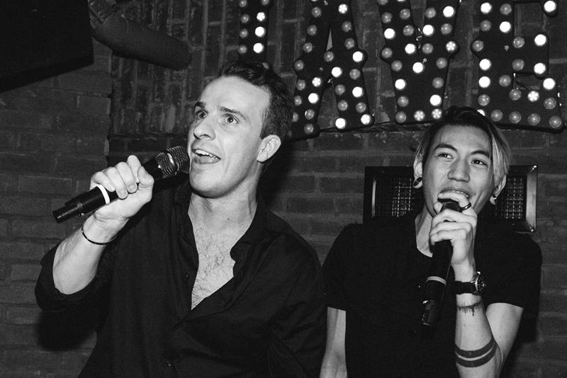 To gjester synger karaoke på en liten scene med mikrofoner og opplyst skilt. Publikum står tett inntil scenen.
