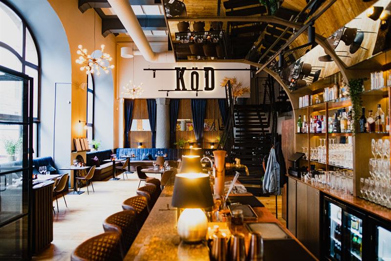 Restaurantinteriør med bardisk, barstoler, hyller med glass og vinflasker, en blå sittegruppe langs veggen og høyt tak