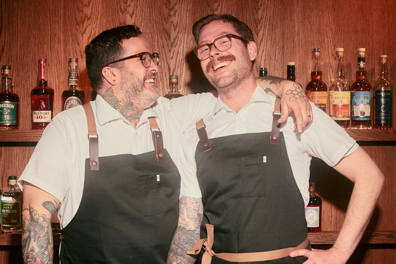 To bartendere smiler bak baren foran hyller med flasker.