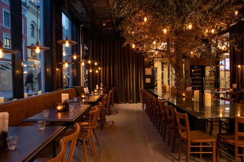 En restaurant med en moderne glassfasade og et varmt, stemningsfullt interiør med trebord, hengende lamper og dekket bord langs vinduene.