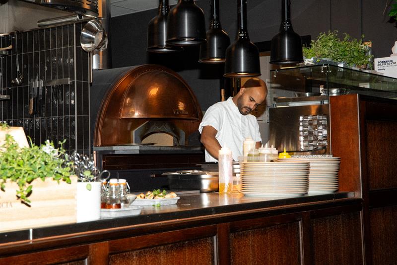 En kokk tilbereder mat ved disken foran en vedfyrt ovn i et moderne restaurantkjøkken.