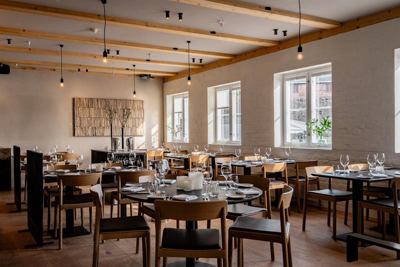 Restaurantinteriør med svarte bord, lyse stoler og lyspærer som henger ned fra taket