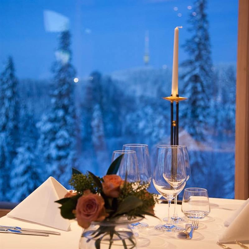 Et elegant dekket bord ved vinduet, med levende lys og et vinterlandskap med skog utenfor.