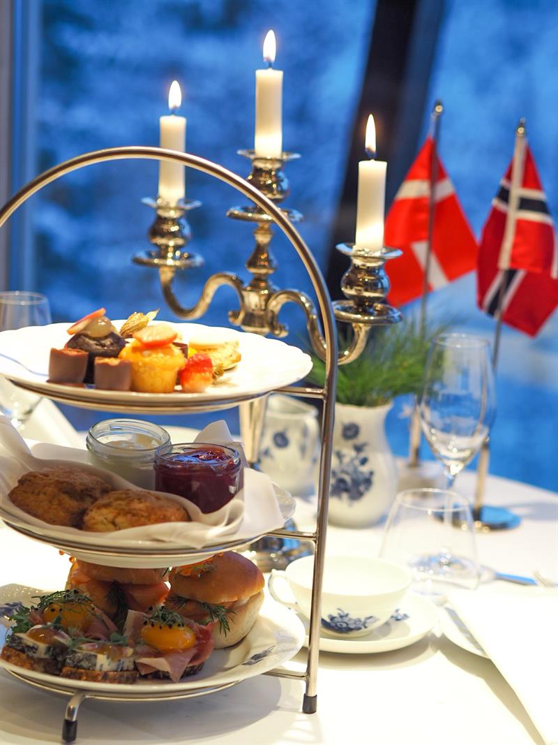 Elegant ettermiddagste servert på et etasjestativ med tente stearinlys og norske flagg ved vinduet.