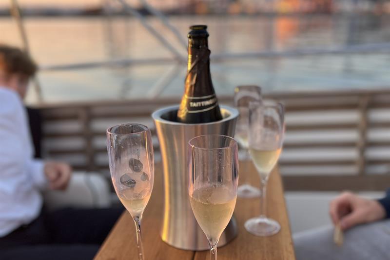 Et glass musserende vin serveres på dekk med utsikt over havnen i solnedgangen.