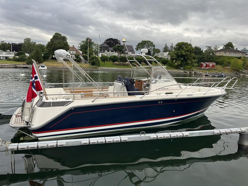En båt ligger fortøyd ved en brygge med hus og hager langs vannkanten.