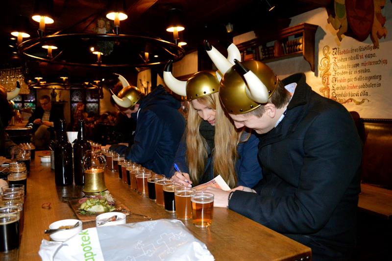 En gruppe mennesker deltar i en ølsmaking innendørs mens de har på seg vikinghjelmer. Flere ølglass står på rekke og rad på et langt bord.