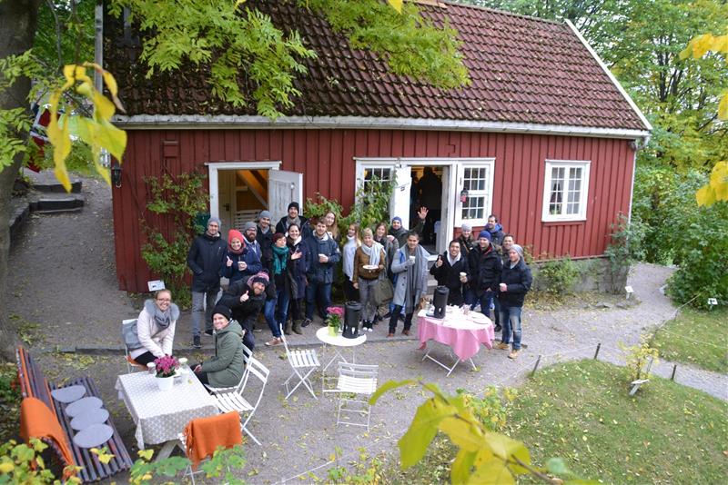 En gruppe mennesker står samlet utenfor en rød trebygning omgitt av natur. Det ser ut til at de er på et sosialt utflukt.