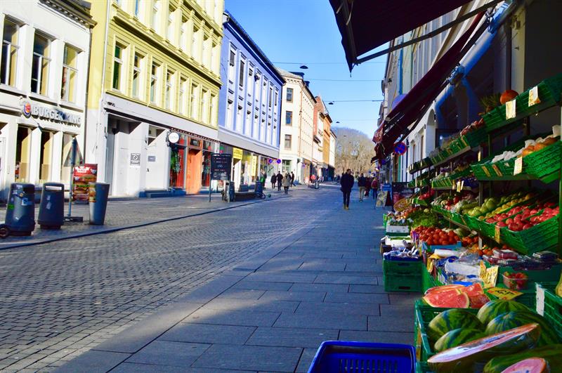 En rolig gate i Oslo med fargerike bygninger og en frukt- og grønnsaksbutikk i forgrunnen. Folk går langs fortauet i solskinnet.