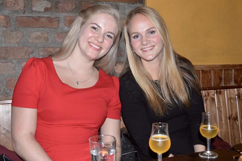 To personer sitter ved et bord og smiler til kameraet, hver med et glass øl i hånden. Stemningen er avslappet og hyggelig.