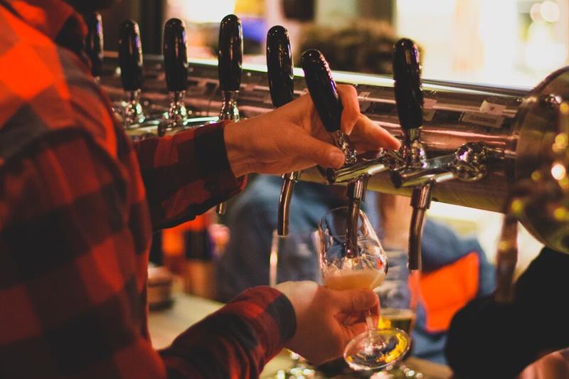 En person skjenker øl fra flere tappekraner i en livlig bar. Stemningen er varm og hyggelig.