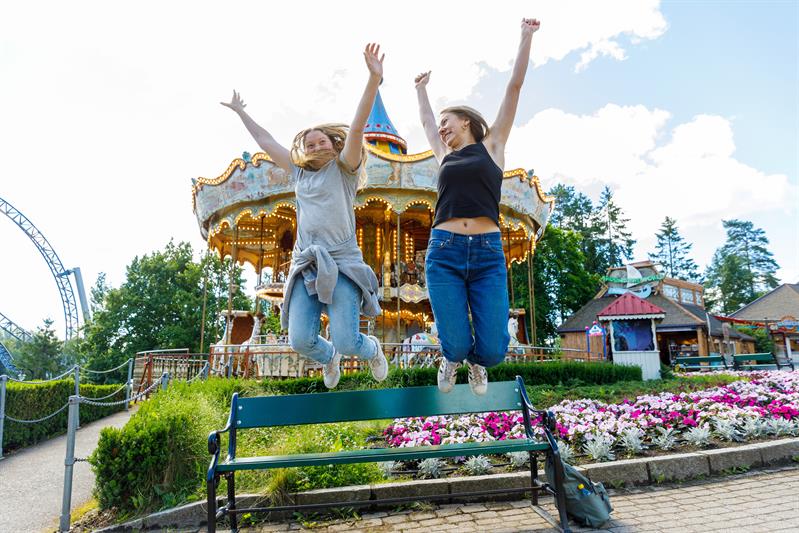Two girls jumping from a park bench in front of a carousel